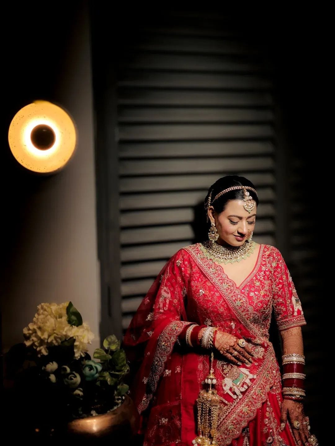The bride in red looking down softly beside dark shutters, lit by a round wall lamp