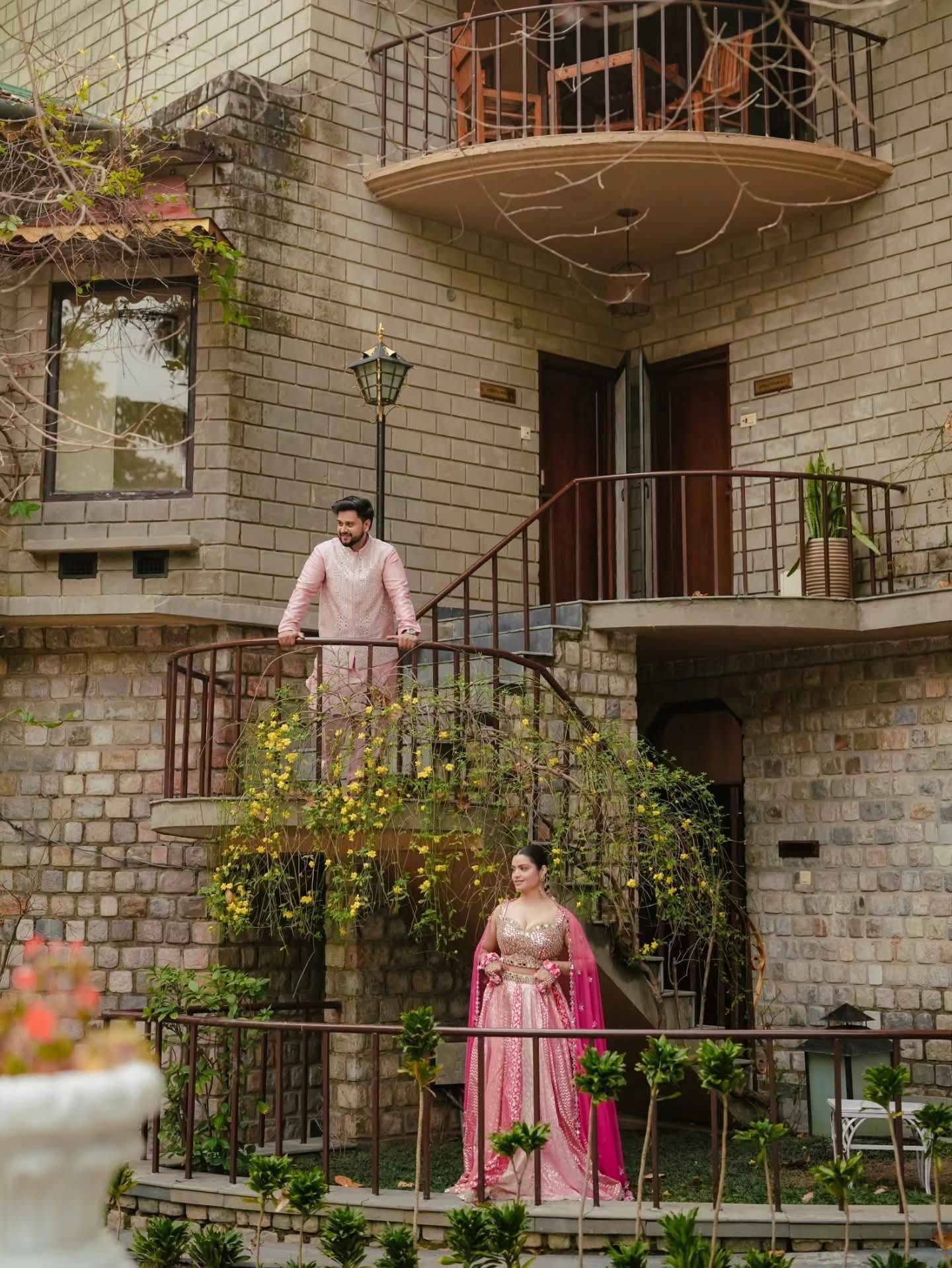 The groom on a flower-draped balcony, the bride in the cottage garden beneath him