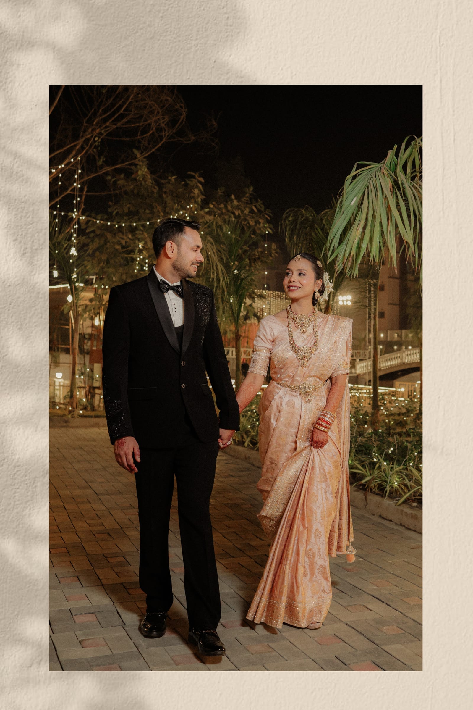 A black tuxedo and a peach silk saree, hand in hand on a fairy-lit garden path