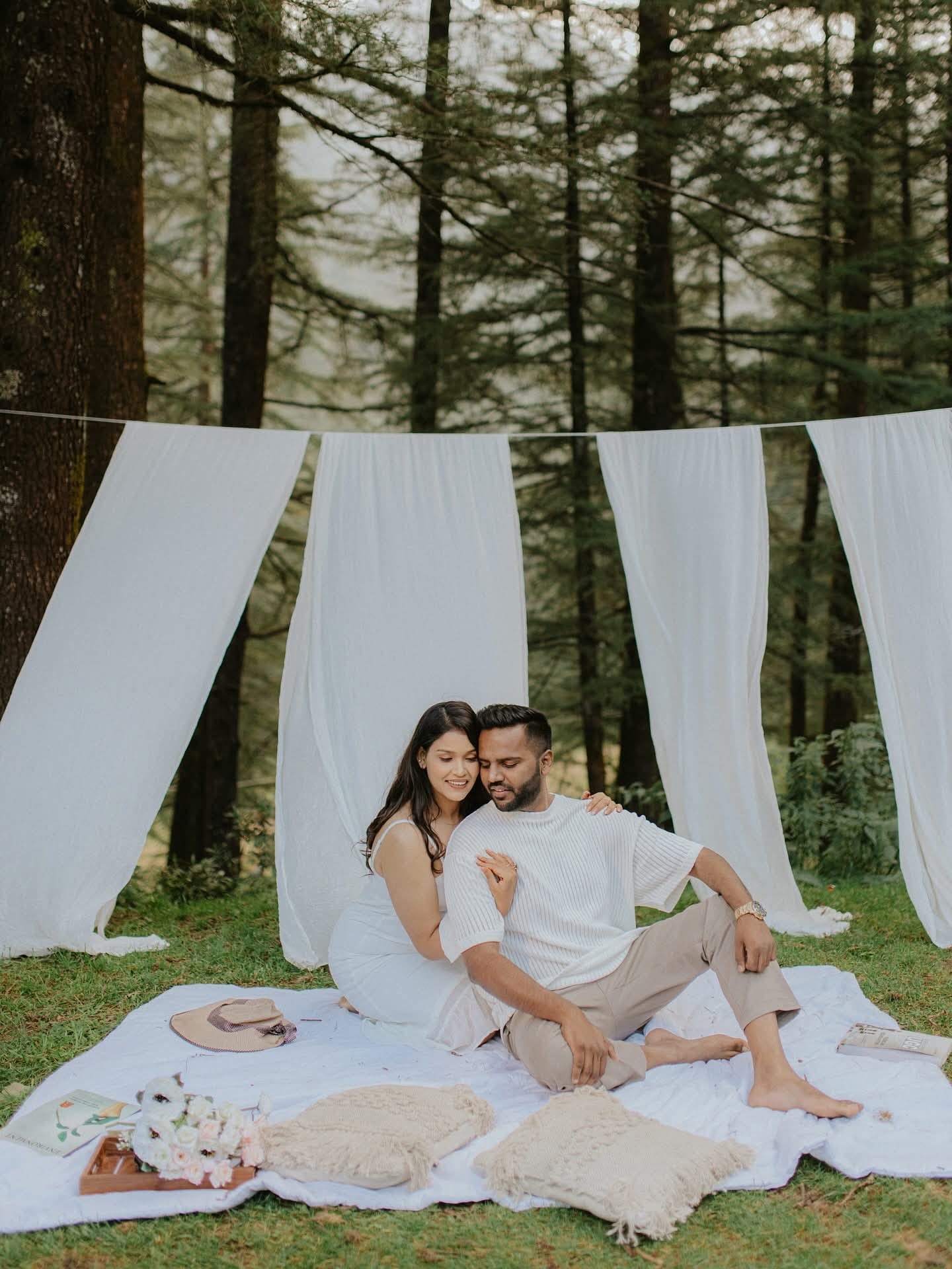 The couple on a picnic blanket beneath white drapes strung between tall pines