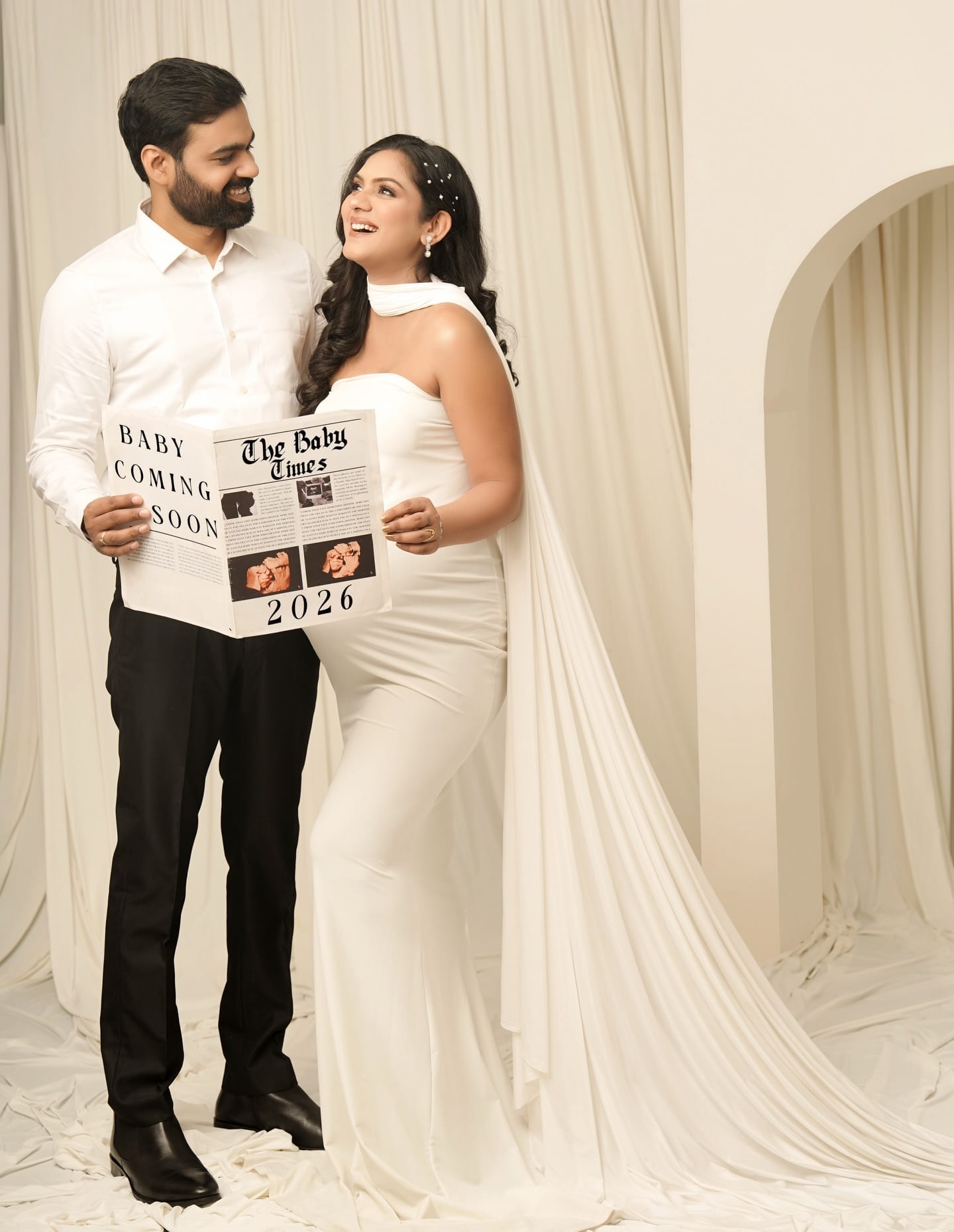 A smiling couple holding a 'Baby Coming Soon' prop newspaper in the studio