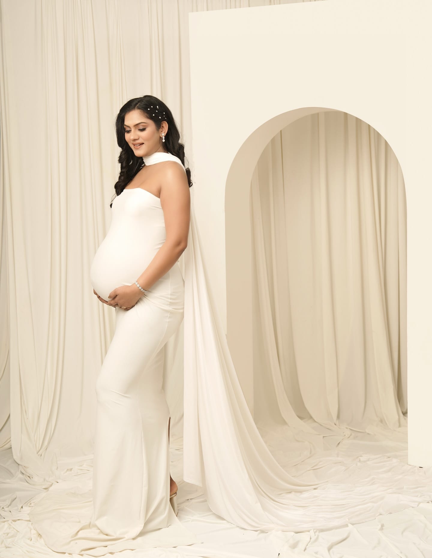 A mother-to-be in a flowing white gown, cradling her bump beside a cream arch
