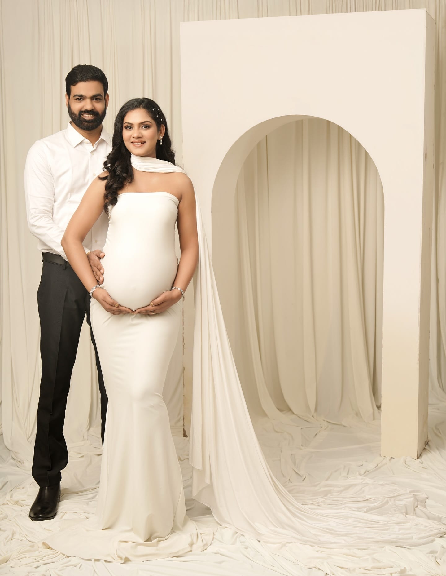 The couple standing together, hands cradling the bump, in the cream studio